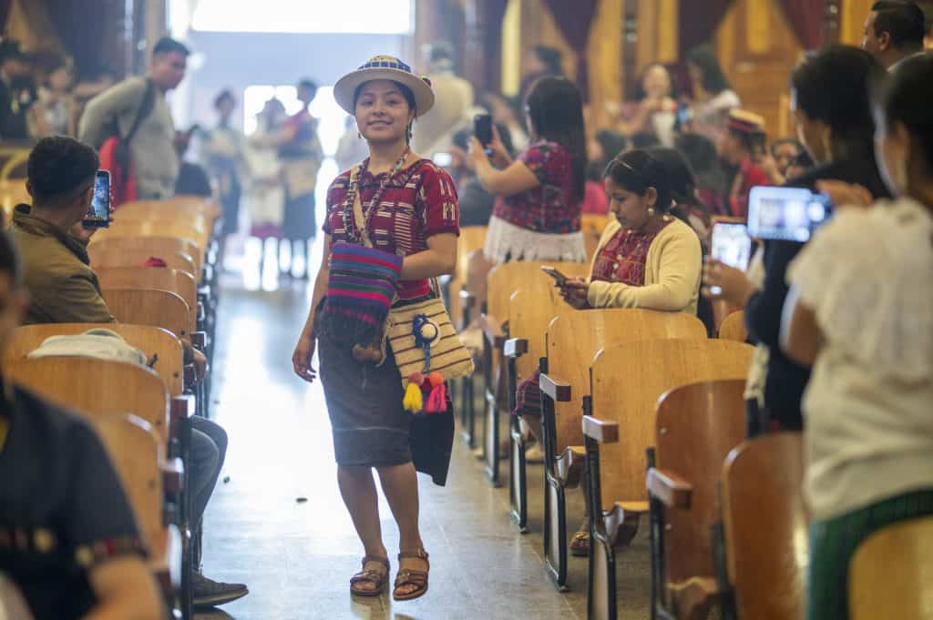 Colorida mujer indígena en vestimenta tradicional, en aeropuerto turístico, esperando traslado o ingreso a un evento cultural. Personas usando celulares capturan momentos en el aeropuerto, reflejando turismo y cultura.
