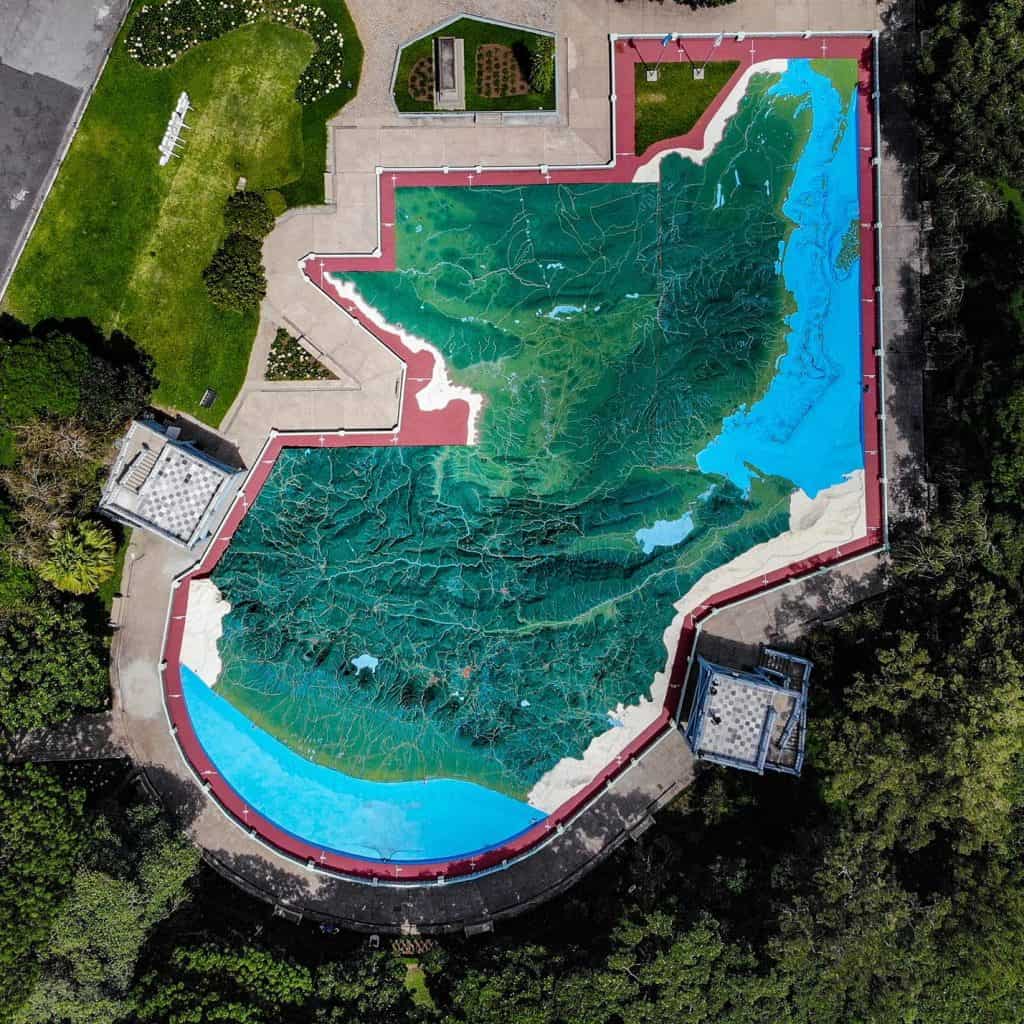 Jardín con mural de mapa topográfico pintado en el suelo, rodeado de áreas verdes y caminos, en un lugar público y cultural, promoviendo la educación ambiental y el arte al aire libre.