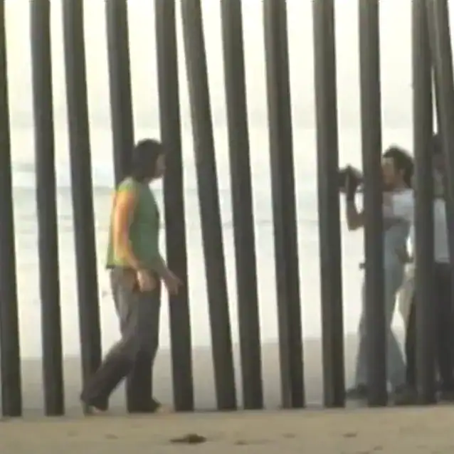 Migrantes frente a barrera de metal en frontera, buscando ingreso o salida, situación común en migración y derechos humanos, en contexto de migrantes en movimiento.