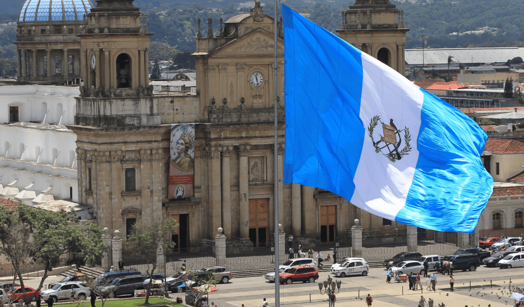 Bandera de Guatemala ondeando frente a una iglesia histórica en la ciudad, símbolo de identidad y orgullo nacional. Publicación en SoyMigrante.com REVISTA, destacando cultura y migración en Guatemala.