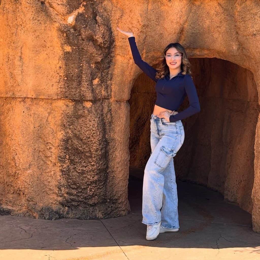 Joven mujer con gafas y jeans anchos en un entorno rocoso, promocionando la revista SoyMigrante.com REVISTA, destacando cultura migrante y fotografía de viaje para comunidad hispanohablante.