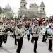Jóvenes músicos militares tocando instrumentos en una plaza concurrida con una multitud y un edificio histórico en el fondo.