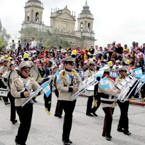 Jóvenes músicos militares tocando instrumentos en una plaza concurrida con una multitud y un edificio histórico en el fondo. – SoyMigrante.com REVISTA