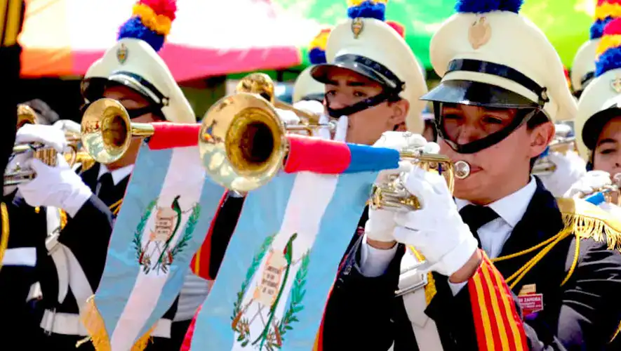 Banda de músicos en uniforme tocando instrumentos en un desfile con banderas de Guatemala en un evento cultural.