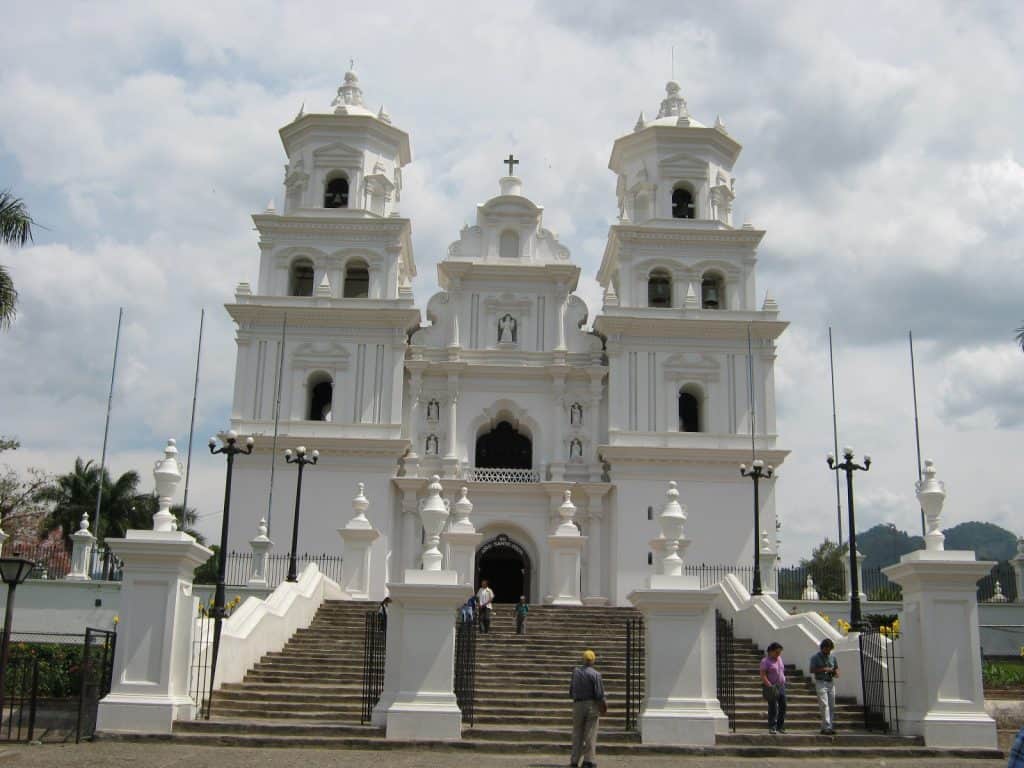 Majestuosa iglesia colonial blanca en un día nublado, con turistas y visitantes en las escalinatas, destacando la arquitectura religiosa y cultural en un destino turístico en México. – SoyMigrante.com REVISTA