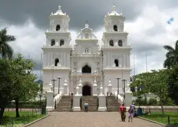 La Basílica de Esquipulas, tesoro colonial de oriente – SoyMigrante.com REVISTA Majestuosa iglesia colonial blanca en un entorno verde, con un cielo nublado, varios visitantes caminando hacia las escaleras principales, resaltando la arquitectura histórica y cultural en un día lluvioso.