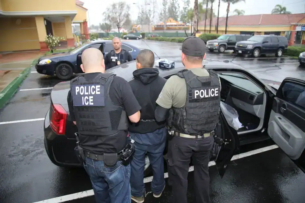Un detenido siendo arrestado por oficiales de policía ICE en un estacionamiento bajo la lluvia, durante una operación de inmigración en Estados Unidos, en presencia de agentes uniformados con chalecos antibalas. – SoyMigrante.com REVISTA