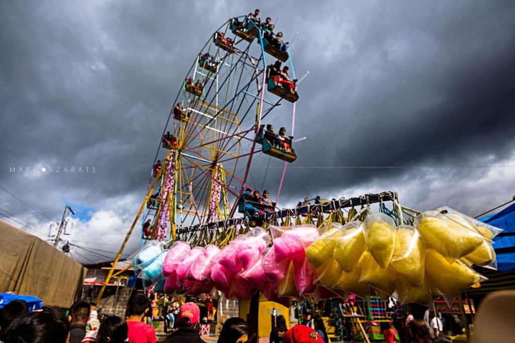 Vuelta en feria con nubes oscuras, atracciones como la rueda de la fortuna y puestos con algodones de azúcar, en un ambiente de celebración y diversión en un lugar popular de migrantes. – SoyMigrante.com REVISTA
