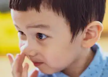 Niño pequeño con expresión curiosa, tocándose la cara, en un entorno educativo, promoviendo temas de infancia y aprendizaje.