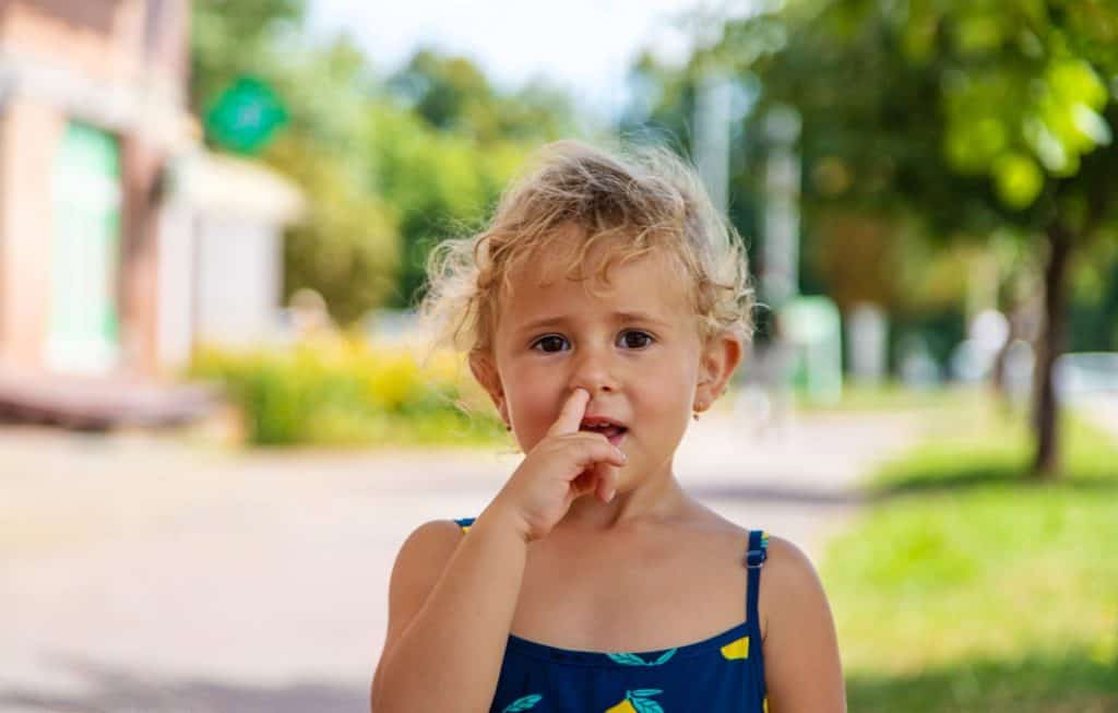 A los niños se les debe enseñar a cuidar de su nariz. (Foto: ENVATO) – SoyMigrante.com REVISTA Niña pequeña en un parque, señalando su nariz con expresión pensativa, en un entorno al aire libre con árboles y senderos, promoviendo bienestar infantil y salud mental para niños. – SoyMigrante.com REVISTA