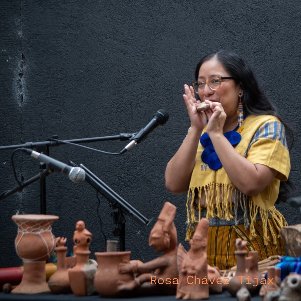 Técnica en música y cultura indígena tocando la flauta mientras presenta cerámica tradicional en evento cultural. – SoyMigrante.com REVISTA