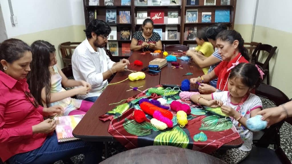 Tejido y manualidades en grupo, actividades culturales para inmigrantes en México, promoción de integración y aprendizaje artístico, taller comunitario, diversidad cultural, mujeres y niños participando en artesanías. – SoyMigrante.com REVISTA