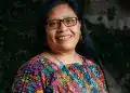 Mujer indígena con gafas, sonriendo y vestida con ropa colorida y patrón tradicional, representando cultura y comunidad en un entorno natural. – SoyMigrante.com REVISTA