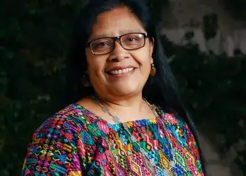 Mujer indígena con gafas, sonriendo y vestida con ropa colorida y patrón tradicional, representando cultura y comunidad en un entorno natural.