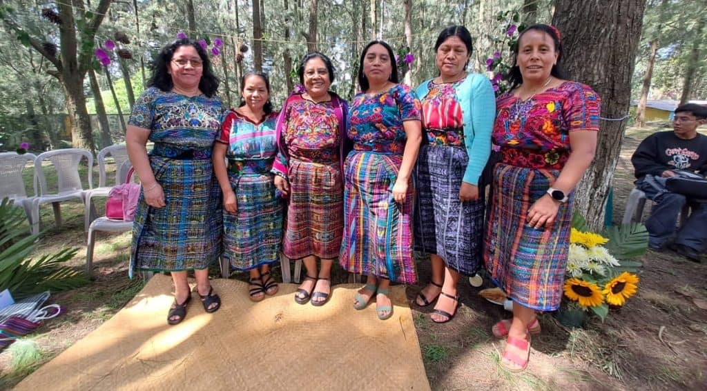 Mujeres indígenas vestidas con trajes tradicionales en un entorno natural, participando en una celebración cultural en la comunidad. – SoyMigrante.com REVISTA