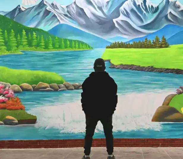 Paisaje pintado de montañas, río y naturaleza con persona observando mural artístico en la revista SoyMigrante.com.
