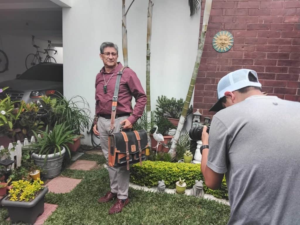Hombre con gafas y chaqueta en una entrevista fotográfica en el jardín de una casa, rodeado de plantas y decoraciones, con un fotógrafo tomando la imagen. – SoyMigrante.com REVISTA