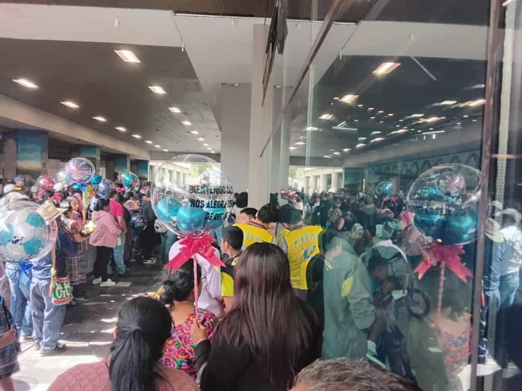 Multitud de migrantes en evento de bienvenida en un aeropuerto, con balloons decorativos, señalando una celebración de retorno y alegría en la comunidad migrante.