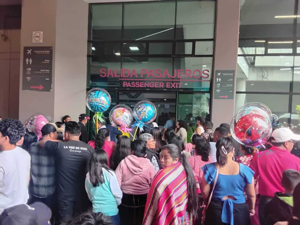 Multitud de pasajeros en la salida de aeropuerto en colorida escena de bienvenida con globos, en el contexto del viaje o migración, ideal para artículos sobre viajes, movilidad y comunidad migrante.