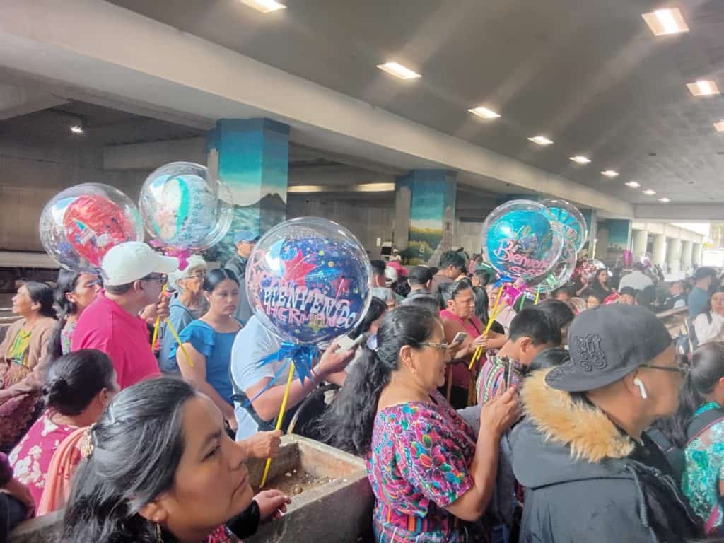 Muchos migrantes en una estación de transporte con globos de bienvenida, celebrando su llegada y reuniéndose en una congregación bajo un puente o estación pública.