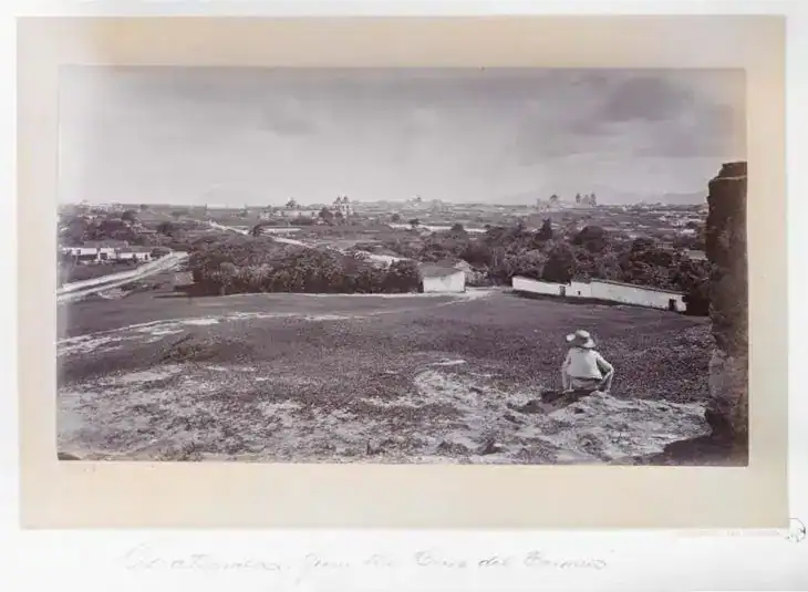 Paisaje rural en blanco y negro con una persona sentada en un campo, vista panorámica de un pueblo con casas y naturaleza, destacando la historia y cultura de los migrantes en México.