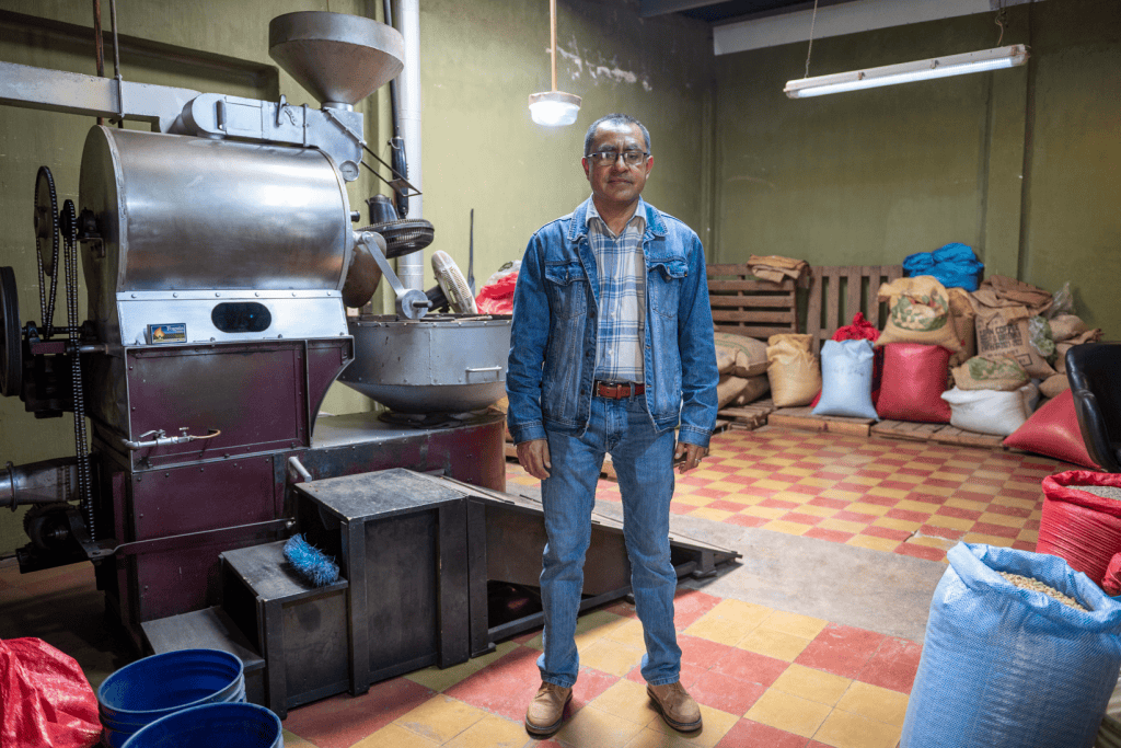 Molino de café industrial en planta de producción con hombre en primer plano en interior de fábrica de café. – SoyMigrante.com REVISTA