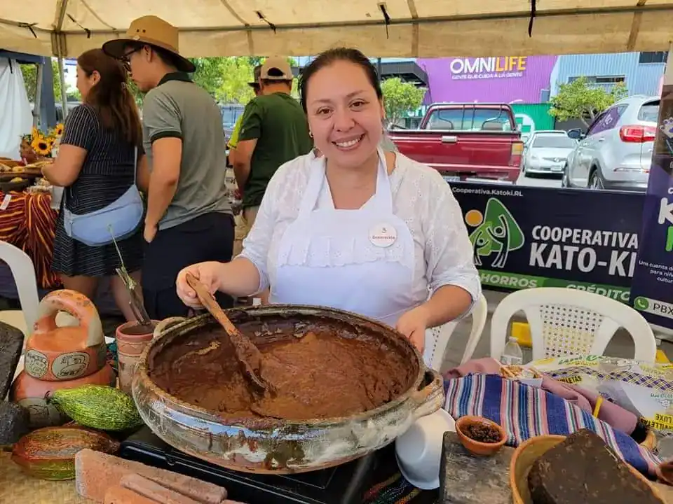 Chile tradicional preparado en una olla grande en un puesto de comida en un evento cultural. Persona sonriendo con delantal, rodeada de productos autóctonos y ambiente festivo. – SoyMigrante.com REVISTA