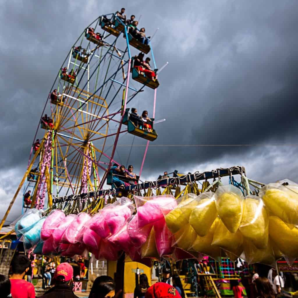 Niños disfrutando en la feria con una noria gigante y puestos con algodón de azúcar, en un día con nubes oscuras, en la revista SoyMigrante.com REVISTA, que habla sobre experiencias y cultura migrante. – SoyMigrante.com REVISTA