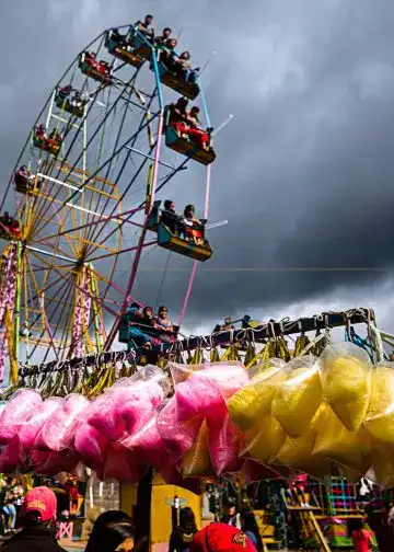Niños disfrutando en la feria con una noria gigante y puestos con algodón de azúcar, en un día con nubes oscuras, en la revista SoyMigrante.com REVISTA, que habla sobre experiencias y cultura migrante.