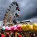 Niños disfrutando en la feria con una noria gigante y puestos con algodón de azúcar, en un día con nubes oscuras, en la revista SoyMigrante.com REVISTA, que habla sobre experiencias y cultura migrante.