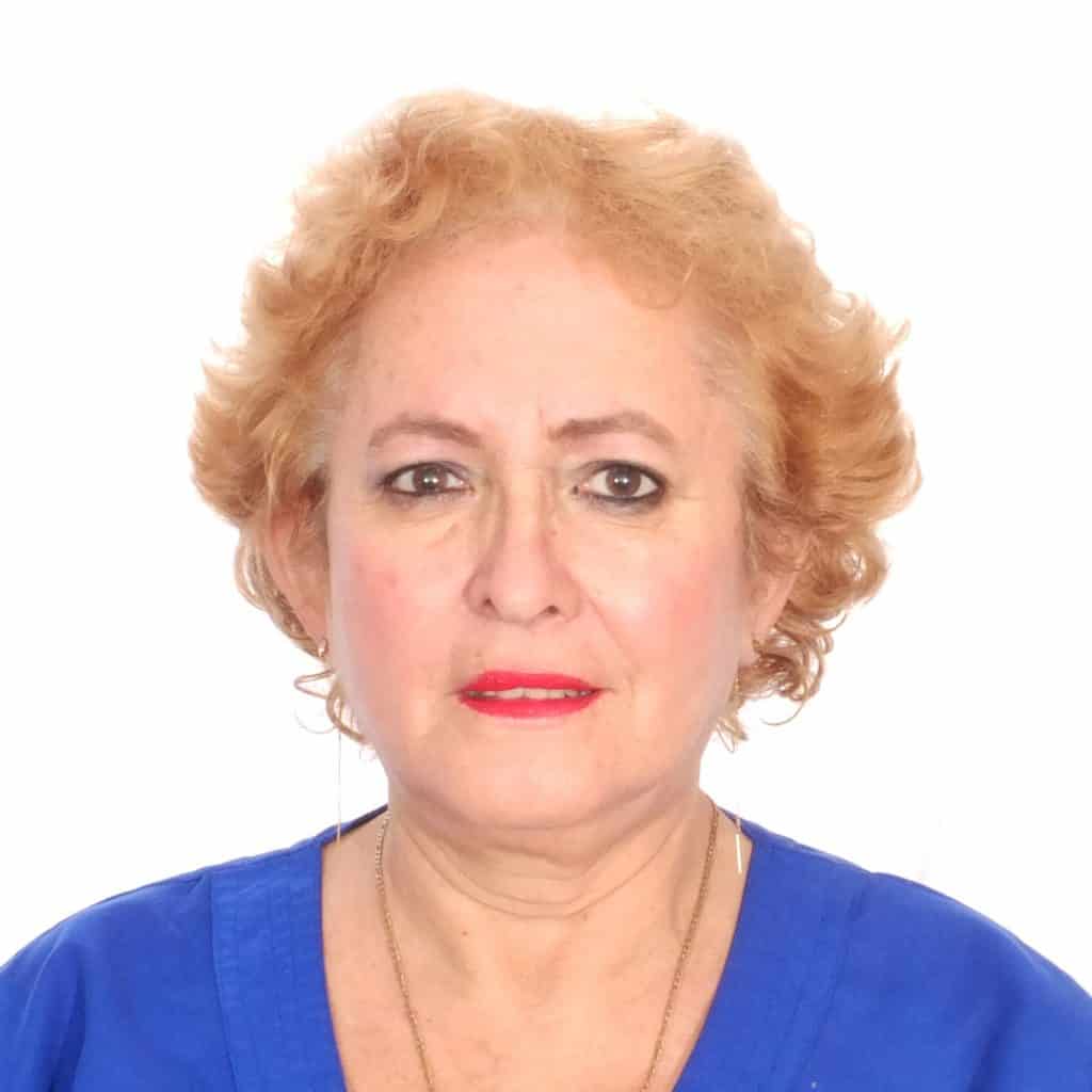 A woman with short, curly blonde hair wearing a blue top and red lipstick, looking directly at the camera against a white background, representing migrant stories and indigenous identity. – SoyMigrante.com REVISTA
