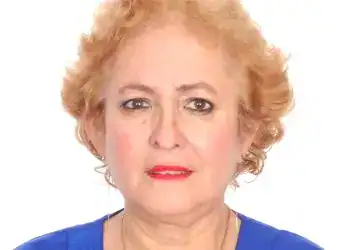 A woman with short, curly blonde hair wearing a blue top and red lipstick, looking directly at the camera against a white background, representing migrant stories and indigenous identity.
