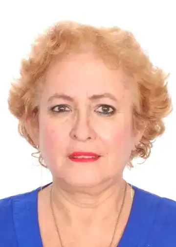 A woman with short, curly blonde hair wearing a blue top and red lipstick, looking directly at the camera against a white background, representing migrant stories and indigenous identity. – SoyMigrante.com REVISTA
