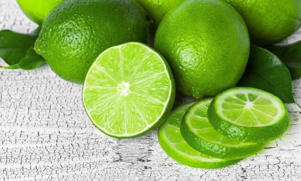 Limas verdes frescas y cortadas en una mesa con textura de madera blanca, destacando su frescura y sabor cítrico, ideales para recetas saludables y bebidas naturales. – SoyMigrante.com REVISTA