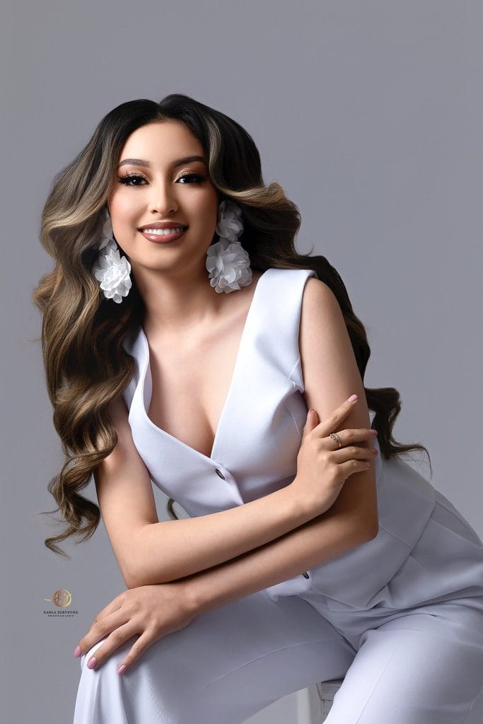 La belleza de Lindsey integra rostro, figura y corazón: una personalidad forjada a partir de desafíos que, sí, le hicieron pasar etapas difíciles, pero que se transformaron en altos ideales. (Fotografía Karla Zertuche, cortesía Lindsey Mazariegos) – SoyMigrante.com REVISTA Hermosa mujer con cabello largo y ondas, vestido blanco elegante y accesorios florales, posando en estudio para revista SoyMigrante.com, reflejando estilo, cultura y diversidad. – SoyMigrante.com REVISTA