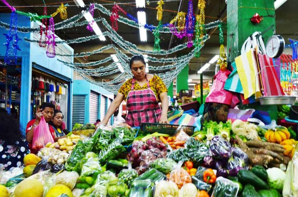 Vendedora en mercado local con frutas y verduras frescas durante temporada navideña, decoraciones festivas y ambiente colorido. – SoyMigrante.com REVISTA