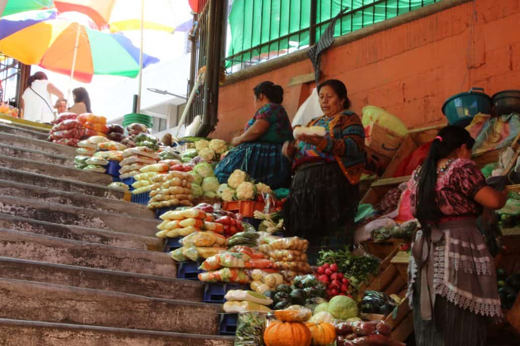Verdulería tradicional en mercado mexicano con frutas y verduras frescas en puestos al aire libre en escalera. Mujeres mexicanas venden productos agrícolas en un ambiente colorido y culturalmente vibrante. – SoyMigrante.com REVISTA