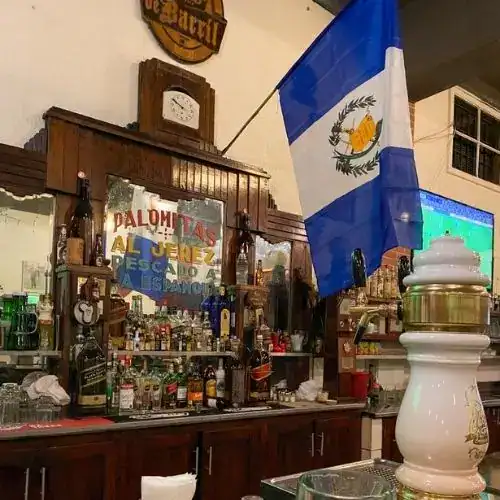 Palmeras y bandera de Guatemala en un bar tradicional con decoración étnica y distintas bebidas en exhibición, transmitiendo ambiente cultural y festivo guatemalteco para la comunidad migrante. – SoyMigrante.com REVISTA
