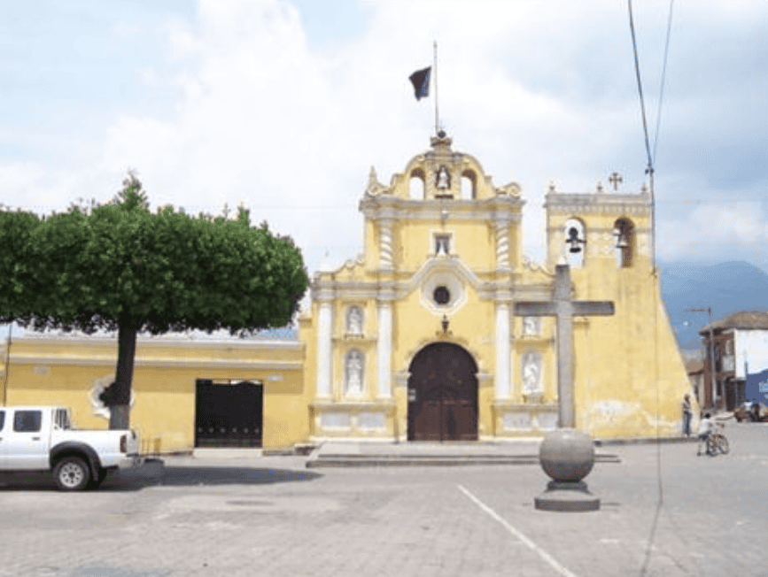 Iglesia colonial amarilla en Guatemala en la plaza principal de un pueblo, con árbol grande a la izquierda, cruz y cielo nublado, representando patrimonio cultural y arquitectura histórica. – SoyMigrante.com REVISTA