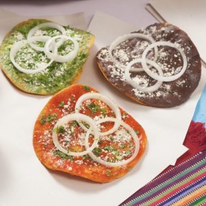 Quesadillas tradicionales con diferentes colores de masa, decoradas con cebolla, cilantro y queso rallado, en un plato con utensilios, representando la gastronomía mexicana en SoyMigrante.com REVISTA.