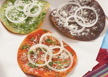 Quesadillas tradicionales con diferentes colores de masa, decoradas con cebolla, cilantro y queso rallado, en un plato con utensilios, representando la gastronomía mexicana en SoyMigrante.com REVISTA.