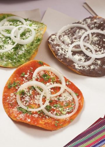 Quesadillas tradicionales con diferentes colores de masa, decoradas con cebolla, cilantro y queso rallado, en un plato con utensilios, representando la gastronomía mexicana en SoyMigrante.com REVISTA.