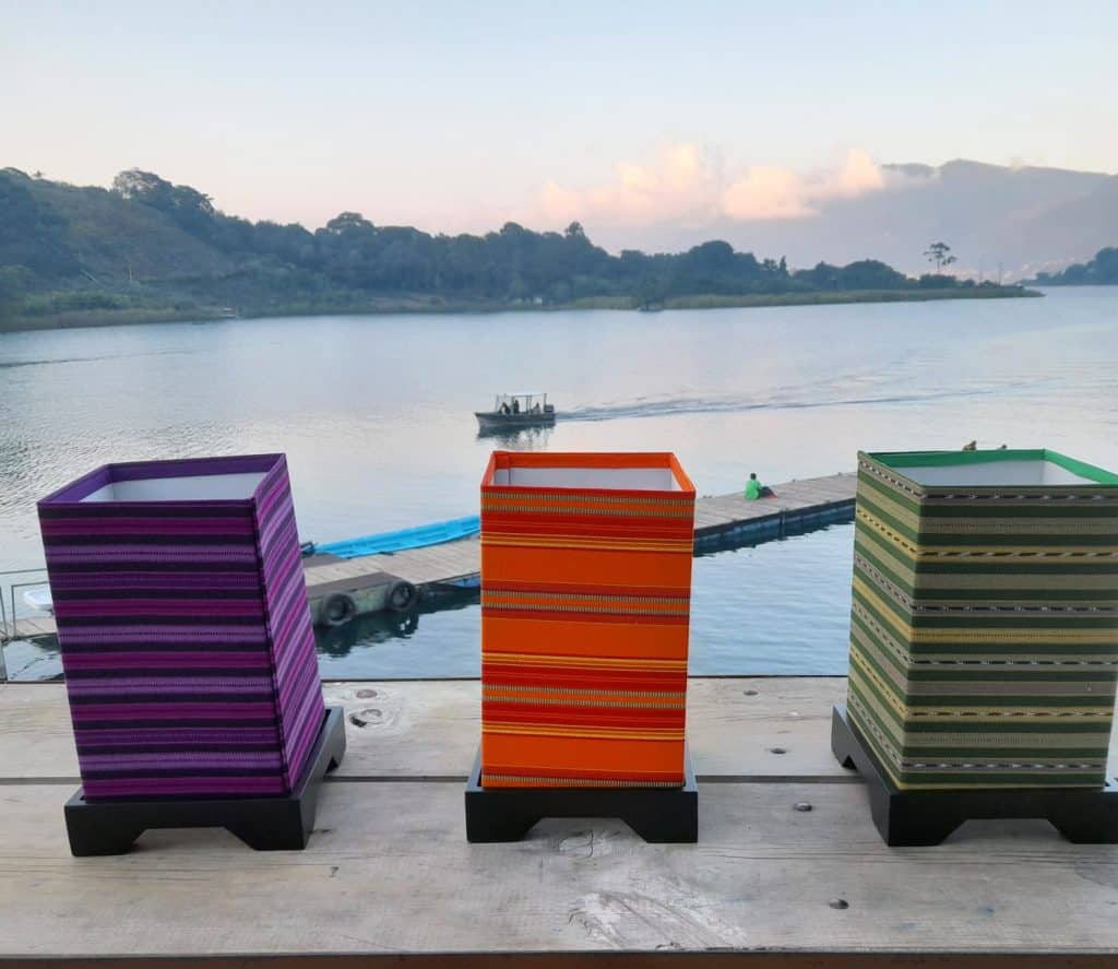 Coloridas sillas de mimbre en un muelle junto a un río, con un bote navegando en el fondo, en un paisaje tranquilo y natural.