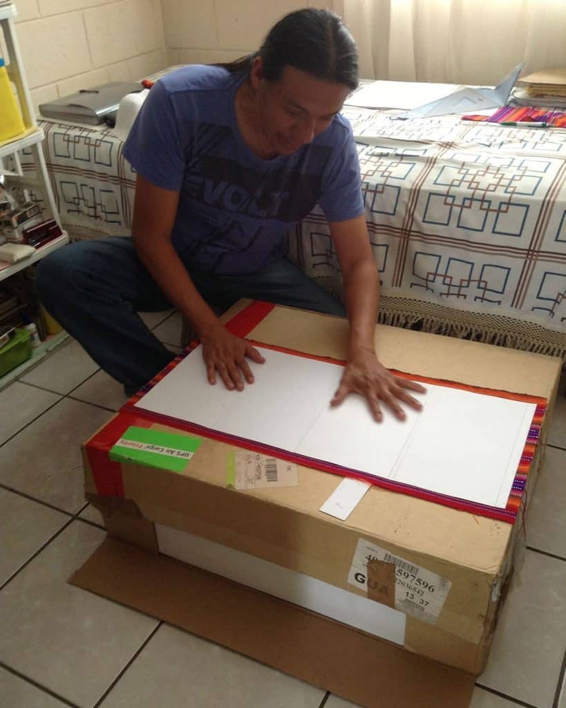 Sobre un hombre que viene del extranjero, revisando una caja de cartón con un libro o cuaderno abierto, en un ambiente interior. – SoyMigrante.com REVISTA