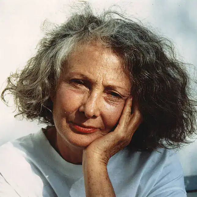 Anciana con cabello gris rizado, expresión pensativa y rostro reflexivo, usando una camiseta blanca, en un entorno natural, simbolizando experiencia y sabiduría. – SoyMigrante.com REVISTA