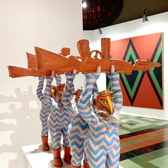 Crianzas disfrazadas con máscaras y trajes a rayas sosteniendo una escopeta de madera en exposición de arte moderno.