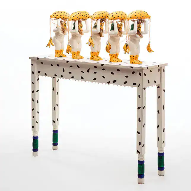 Niños de juguete en un mesa blanca con manchas negras, sosteniendo paraguas con patucos y ropa a rayas, en una imagen creativa y colorida.