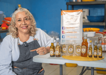 Aceite artesanal y productos naturales en la feria gastronómica de SoyMigrante.com REVISTA, con mujer emprendedora mostrando ingredientes y aceites en un ambiente de cocina o mercado.