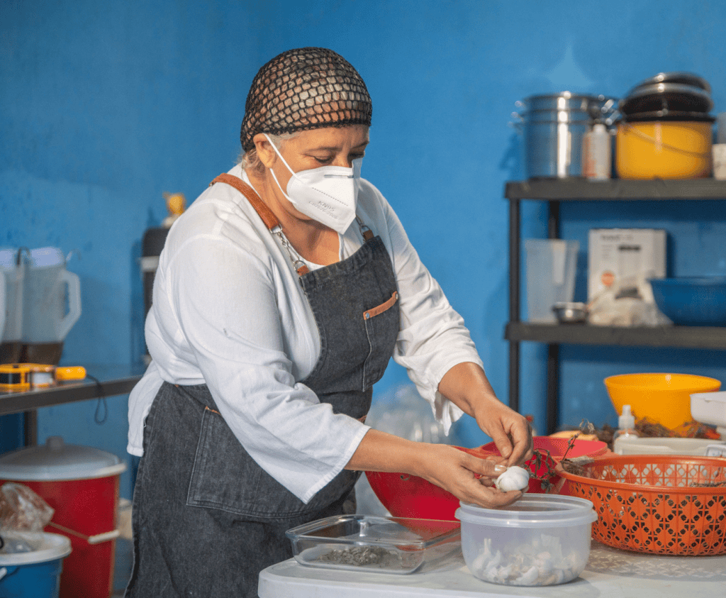 Artesana preparando comida en cocina, con mascarilla y delantal, en un ambiente de cocina casera. – SoyMigrante.com REVISTA