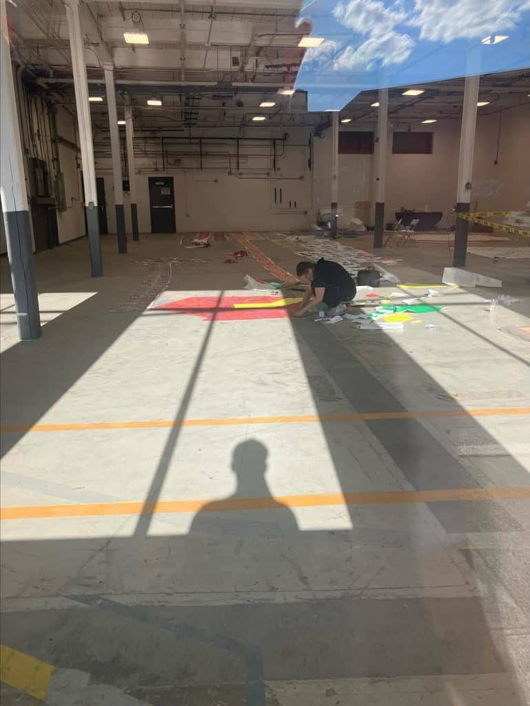 La artista Magda León escribió acerca de esta fotografía: "Mi postura quizá no sea la mejor, pero sin querer se capta el cielo sobre mí. Así me siento cuando estoy creando: como si tocara un pedacito de Dios. Me pierdo en el papel, en los colores, en las formas; el tiempo deja de existir. Cada recorte lo posiciono con cuidado, para que pueda contar las historias de los que vivimos lejos, cerca y entremedio. Amo lo que hago y lo veo como un regalo del que me siento agradecida y muy afortunada de tener. Por la niña de 9 años que emigró y que siempre soñó con ser artista. La abrazo y le digo, sí se pudo." (Foto cortesía Magda León) – SoyMigrante.com REVISTA